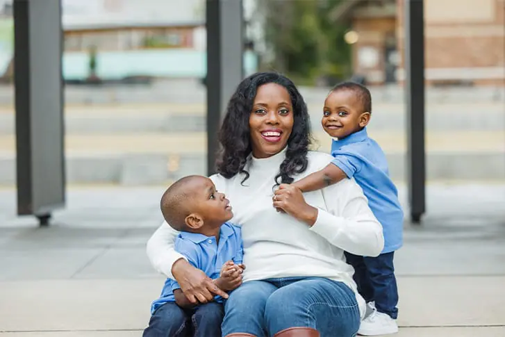 A woman sits on a street curb and smiles at the camera. She is hugging wwo preschool-aged children on either side of her.