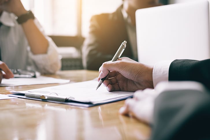 Man sits at a table with other people and writes notes on a legal pad.